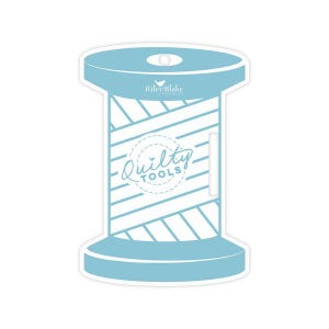May include: A light blue sticker in the shape of a spool of thread, with the words "Quilty Tools" in a circular design. The brand name "Riley Blake Designs" is printed at the top. The spool has a white and blue striped pattern.