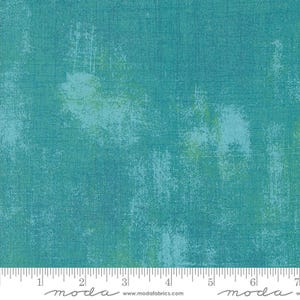 May include: A teal blue fabric with a mottled, textured surface. The fabric has a subtle green and blue color variation. The fabric is measured in inches with a ruler that says "moda" on it.