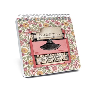 May include: A spiral-bound notepad with a floral pattern in pink, yellow, and green. The notepad features a pink typewriter illustration with the word "Notes..." printed on the paper.