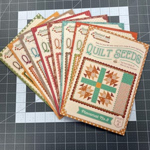 May include: A collection of eight rectangular quilt pattern booklets titled Quilt Seeds by Lori Holt of Bee in my Bonnet. Each booklet features a unique quilt block design, decorative borders, and the text Baked with Love on a cream background.
