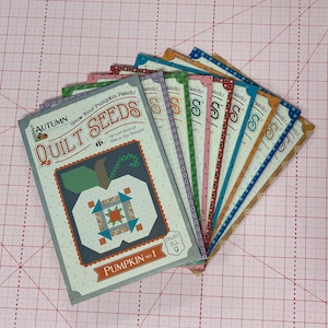 May include: A collection of nine quilt seed packets, each featuring a unique design. The packets are labeled "Autumn Quilt Seeds" and "Pumpkin No. 1." The packets have a pumpkin design and are arranged on a pink grid surface.