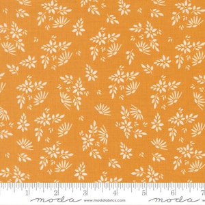 May include: A mustard yellow fabric featuring a repeating pattern of small white flowers and delicate leafy sprigs. A white ruler is visible at the bottom edge, showing measurements in inches, with the text moda and www.modafabrics.com printed.