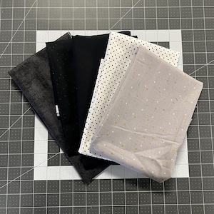 May include: A collection of folded fabric pieces in various patterns and colors. The fabrics include solid black, black with small white polka dots, and a light gray fabric with gold star patterns. Other fabrics are dark gray and black.
