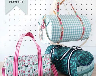 May include: Three different styles of lined duffel bags. The bags are made of fabric with various patterns, including a blue and white gingham, a nautical flag pattern, and a teal and white whale pattern. The bags have pink, white, and black straps. The bags are hanging on a wooden rack against a white wall with black polka dots.