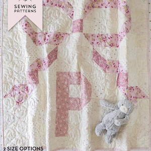 May include: A white and pink quilt with a bow and the letter "P" design. The quilt features floral patterns and is accompanied by a gray stuffed animal. The text on the image reads "Bows & Monograms Quilt".