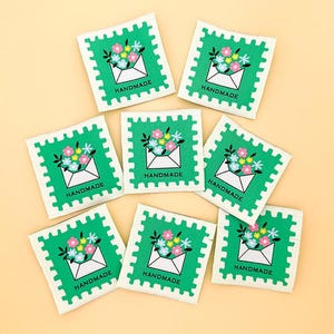 May include: A collection of square fabric labels with a green border, featuring a white envelope overflowing with colorful flowers. The word "HANDMADE" is printed in black at the bottom of each label. The labels are arranged on a light orange background.