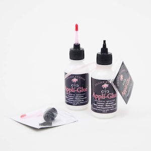 May include: Two bottles of white glue with black caps and red droppers. The bottles are labeled "Appli-Glue Archival Quality" and feature a pink flower logo. The glue is acid-free, pH neutral, water soluble, water reversible, and has an anti-clog precision tip. The bottles are 2 oz each.