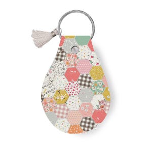 May include: A pink, green, and white patchwork fabric keychain with a silver key ring and a small white tassel. The fabric is patterned with flowers, stars, and geometric shapes. The keychain has the text "Best of You, with You, for You" printed on it.