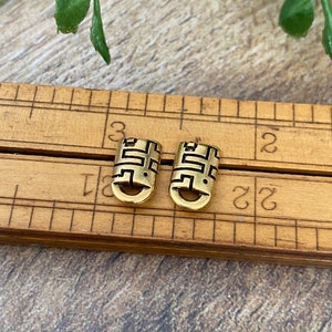 May include: Two gold-colored metal charms with a geometric design. The charms are about 1 cm in length and are sitting on a wooden ruler.