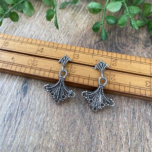 May include: Two silver-toned metal earring findings with a decorative design. The findings are shown on a wooden ruler with markings in inches.