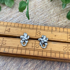 May include: Two silver-toned flower-shaped metal clasps with a loop on the bottom. The clasps are on a wooden ruler with markings.