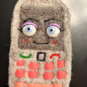 May include: A handmade felt phone-shaped keychain with a cartoon face. The gray phone has a keypad with coral-colored buttons, a green call button, and a red end call button. The phone has blue eyes, purple eyebrows, and a smiling mouth. A blue carabiner clip is attached.