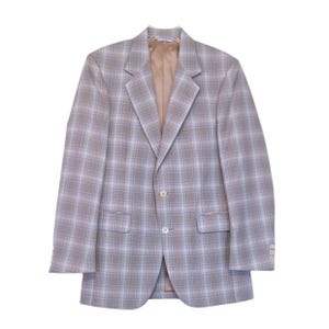 Puede incluir: Una blazer a cuadros marrón claro con cierre de dos botones, solapas con muescas y dos bolsillos delanteros. La blazer tiene un forro de color beige claro y se muestra sobre un fondo blanco. El patrón de cuadros incluye líneas azules claras y rosas.
