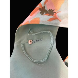 May include: A close-up of a light teal garment with a floral pattern in orange, pink, and brown. The garment has a button closure and a delicate, braided cord detail. The background is black.