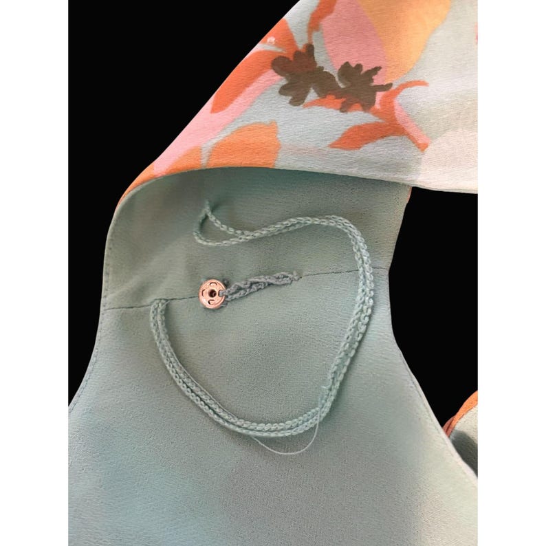 May include: A close-up of a light teal garment with a floral pattern in orange, pink, and brown. The garment has a button closure and a delicate, braided cord detail. The background is black.