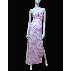 May include: A strapless, floor-length dress in white with a pink floral pattern. The dress features a bow detail at the bust and a high slit on one side. The dress is displayed on a mannequin.