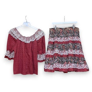 May include: A two-piece vintage outfit featuring a red blouse and skirt. The blouse has a square neckline and lace trim. The skirt is tiered with a floral pattern and lace accents. Both pieces are displayed on hangers.