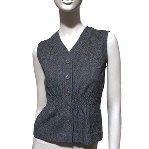 May include: A gray sleeveless vest with a V-neckline and a row of five round buttons down the front. The vest has a cinched waist and is made of a textured fabric.