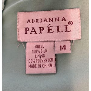 May include: Close-up of a label from Adrianna Papell. The label is rectangular with the brand name in a serif font. The label indicates the garment is size 14, with a shell made of 100% silk and a lining of 100% polyester. The label is on a light blue fabric.