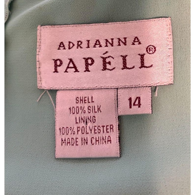 May include: Close-up of a label from Adrianna Papell. The label is rectangular with the brand name in a serif font. The label indicates the garment is size 14, with a shell made of 100% silk and a lining of 100% polyester. The label is on a light blue fabric.