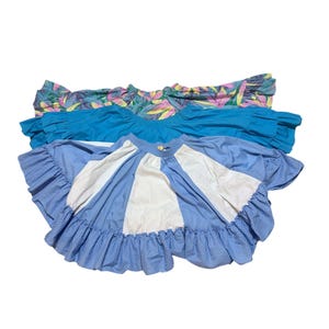 May include: Three colorful skirts. The top skirt has a tropical floral print in shades of pink, green, and yellow. The middle skirt is solid turquoise. The bottom skirt is light blue with white panels and a polka dot pattern, finished with a ruffled hem.