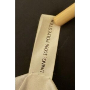 Puede incluir: Primer plano de una etiqueta de tela blanca con el texto "LINING: 100% POLYESTER" impreso en negro. La etiqueta est&aacute; sujeta por un objeto de color claro sobre un fondo oscuro.