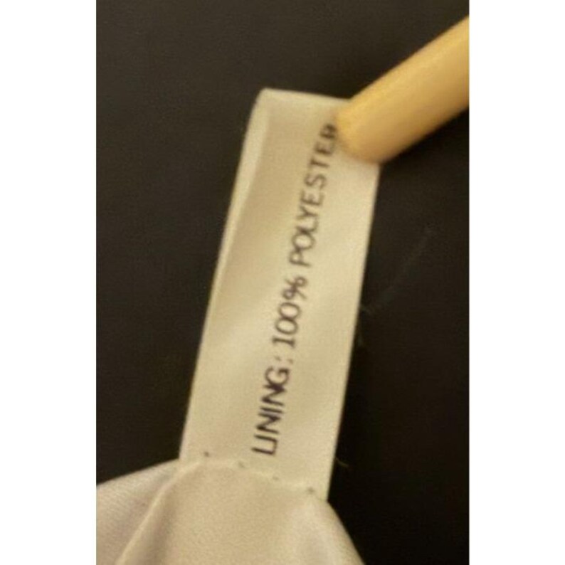 Puede incluir: Primer plano de una etiqueta de tela blanca con el texto "LINING: 100% POLYESTER" impreso en negro. La etiqueta est&aacute; sujeta por un objeto de color claro sobre un fondo oscuro.