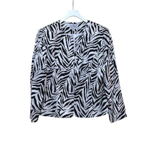 Puede incluir: Chaqueta con estampado de cebra en blanco y negro con cierre de botones. La chaqueta est&aacute; colgada de una percha blanca sobre un fondo blanco. La chaqueta tiene mangas largas y un dise&ntilde;o sin cuello.