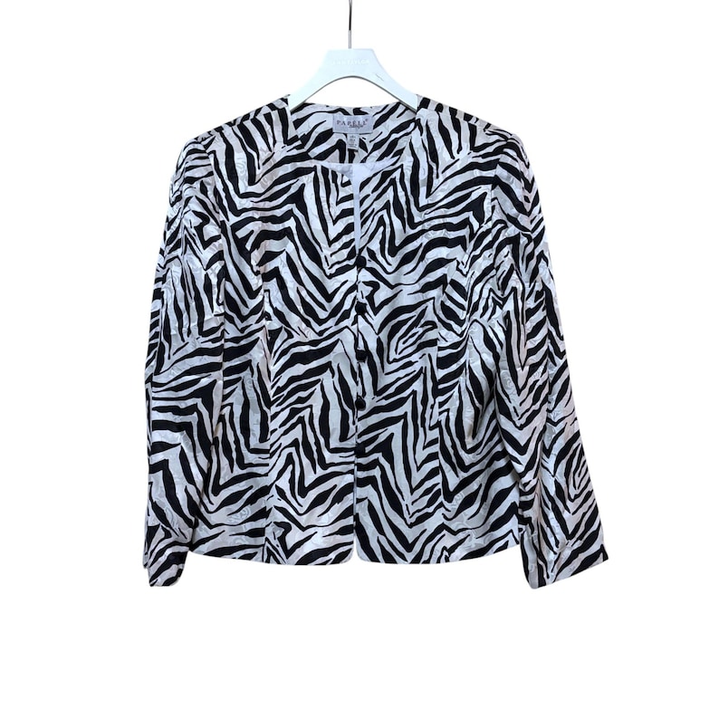 Puede incluir: Chaqueta con estampado de cebra en blanco y negro con cierre de botones. La chaqueta est&aacute; colgada de una percha blanca sobre un fondo blanco. La chaqueta tiene mangas largas y un dise&ntilde;o sin cuello.