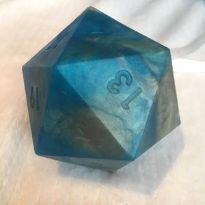 May include: A twenty-sided die featuring a translucent blue and gold marbled resin finish. The die has sharp edges and clearly engraved black numbers, including the number thirteen prominently displayed on the front face and nineteen on the side.