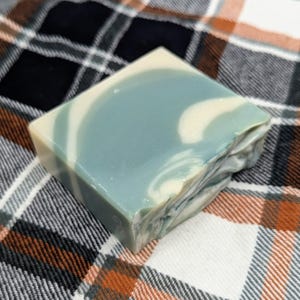 May include: A square bar of handmade soap with a marbled design. The soap features a blend of blue and off-white colors, with a textured, uneven edge on one side. The soap is set against a plaid fabric background.