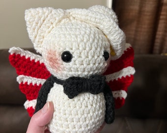 May include: A handmade crochet doll with a white body, head, and ears. The doll has black eyes, rosy cheeks, and a black bow tie. It features red and white striped wings and black arms.