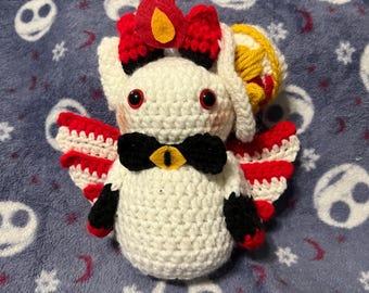 May include: White crocheted plush toy with red and black accents. The toy has a red bow, black eyes, and a yellow bow tie. The toy is wearing a red and white crocheted cape and has a red felt flame on its head.