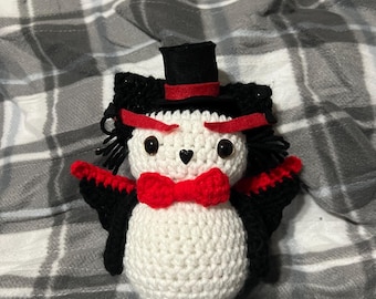 May include: A white crocheted vampire plush toy wearing a black top hat with a red band, a red bow tie, and black wings. The toy has black eyes and a small black heart on its chest.
