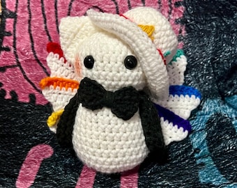 May include: A handmade crochet doll with a white body, black bow tie, and black arms. The doll has a white hat with colorful accents and wings with rainbow colors. The doll is on a pink and black patterned surface.