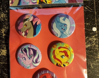 Op de afbeelding: Set van zes ronde knopen met cartoon pony-personages uit de show My Little Pony: Friendship is Magic. De personages zijn Twilight Sparkle, Fluttershy, Rainbow Dash, Pinkie Pie, Applejack en Rarity. De knopen zijn allemaal in verschillende kleuren en hebben een glanzende afwerking. De knopen zijn verpakt in een doorzichtige plastic zak met een rode achterkant waarop "Reformed" staat.