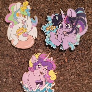 Peut inclure: Trois épingles en émail représentant des poneys de dessin animé dans des scènes estivales. La première épingle montre un poney blanc avec une bouée gonflable rose et jaune, portant des lunettes de soleil et buvant dans un verre. La deuxième épingle montre un poney violet avec une queue rose et bleue, portant un tuba et tenant un poisson. La troisième épingle montre un poney violet avec une queue rose et jaune, portant un maillot de bain et nageant dans l'eau.
