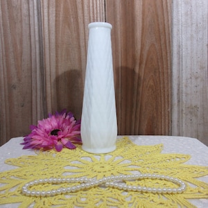 May include: A tall, white, textured vase with a diamond pattern. A pink flower and a strand of pearls are on a yellow doily. The background is a wooden wall.