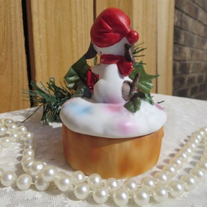 May include: A white ceramic Christmas trinket box with a small dog figurine wearing a red hat and scarf. The dog is sitting on a bed of white snow with green holly leaves. The box is decorated with pink, blue, and white paint.
