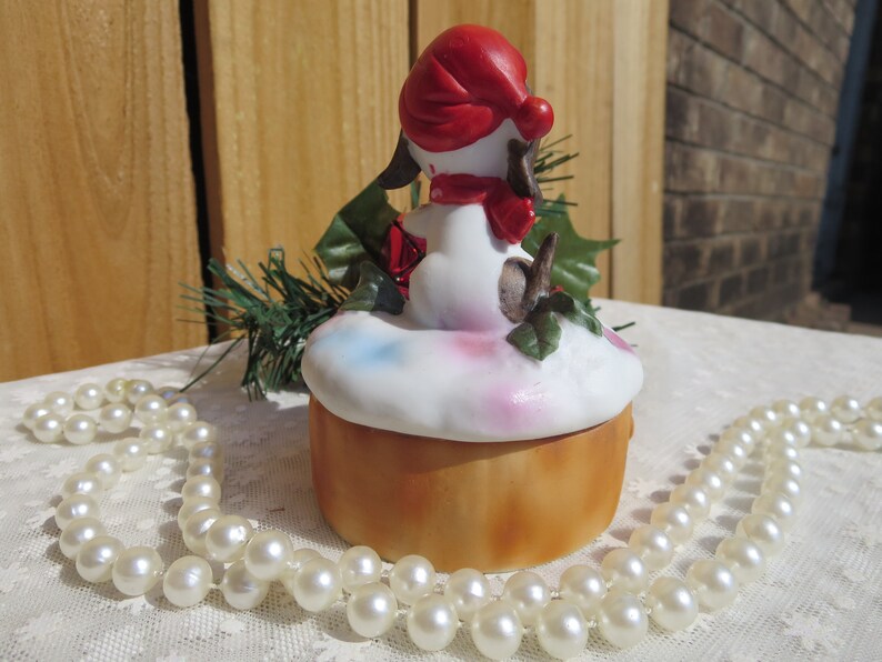 May include: A white ceramic Christmas trinket box with a small dog figurine wearing a red hat and scarf. The dog is sitting on a bed of white snow with green holly leaves. The box is decorated with pink, blue, and white paint.
