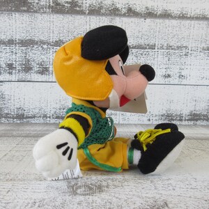 May include: A plush toy of a cartoon character wearing a yellow football helmet, green and yellow jersey, and yellow pants. The character has black ears, a black nose, and white gloves. The toy is sitting on a white wooden surface.