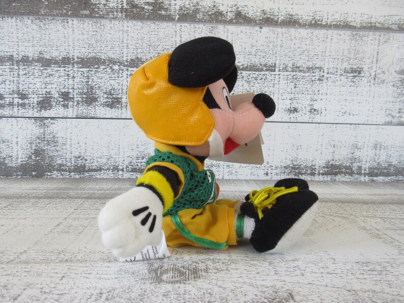 May include: A plush toy of a cartoon character wearing a yellow football helmet, green and yellow jersey, and yellow pants. The character has black ears, a black nose, and white gloves. The toy is sitting on a white wooden surface.