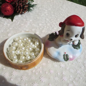 May include: A white porcelain figurine of a dog wearing a red Santa hat. The dog is holding a sprig of holly in its paw. The figurine is sitting on a white base with a blue and pink design. The figurine is in a closed, round, brown and white ceramic box with a white interior. The box is filled with a string of white pearls.
