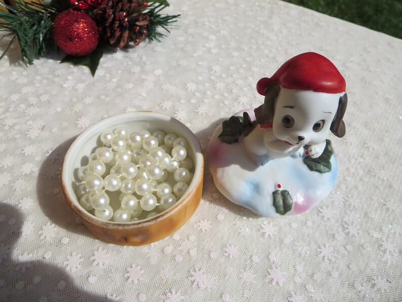 May include: A white porcelain figurine of a dog wearing a red Santa hat. The dog is holding a sprig of holly in its paw. The figurine is sitting on a white base with a blue and pink design. The figurine is in a closed, round, brown and white ceramic box with a white interior. The box is filled with a string of white pearls.