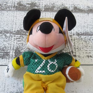 May include: A Mickey Mouse plush toy wearing a green and yellow football uniform with the number 20. The toy has black ears, a red mouth, and is holding a brown football. The background is a white and gray wooden surface.
