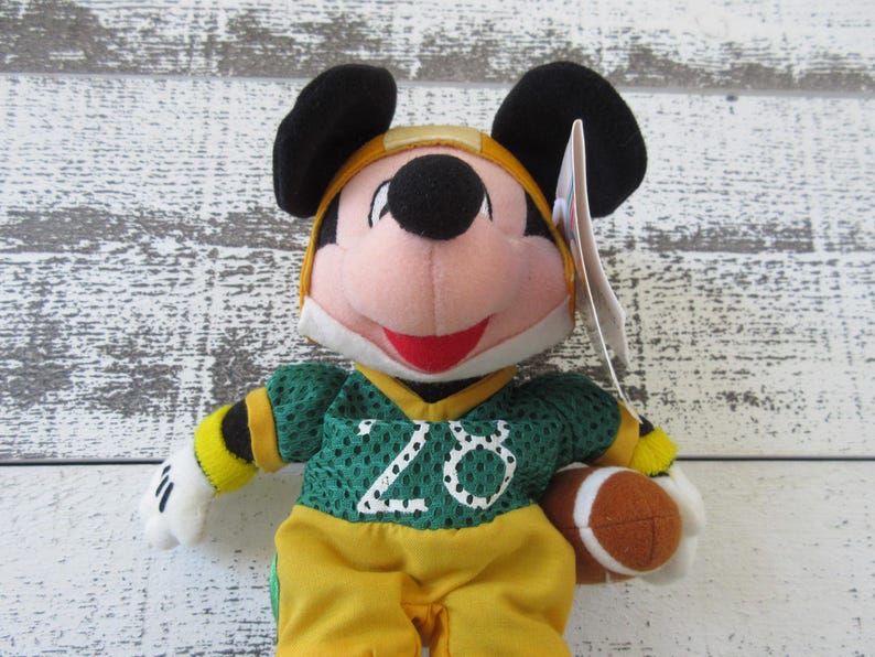 May include: A Mickey Mouse plush toy wearing a green and yellow football uniform with the number 20. The toy has black ears, a red mouth, and is holding a brown football. The background is a white and gray wooden surface.