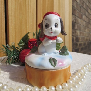 May include: A white porcelain figurine of a dog wearing a red Santa hat and scarf, sitting on a snow-covered base with holly leaves. The base is a brown log-shaped box.