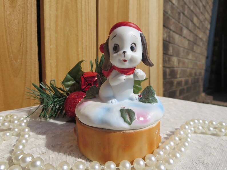 May include: A white porcelain figurine of a dog wearing a red Santa hat and scarf, sitting on a snow-covered base with holly leaves. The base is a brown log-shaped box.