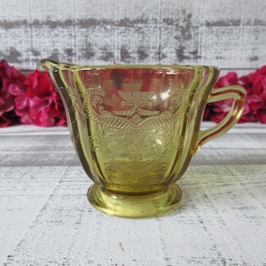 May include: A yellow glass creamer with an ornate design. The creamer has a small handle and a rounded base.