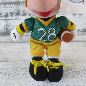 May include: A plush toy of a character dressed in a green and yellow football uniform, holding a brown football. The jersey has the number 28 in white. The character wears black shoes with yellow laces.